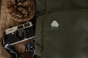 Acrylic pin in the shape of a paw designed with trees and mountains within the paw pinned to a green backpack with a camera and pinecone close by.