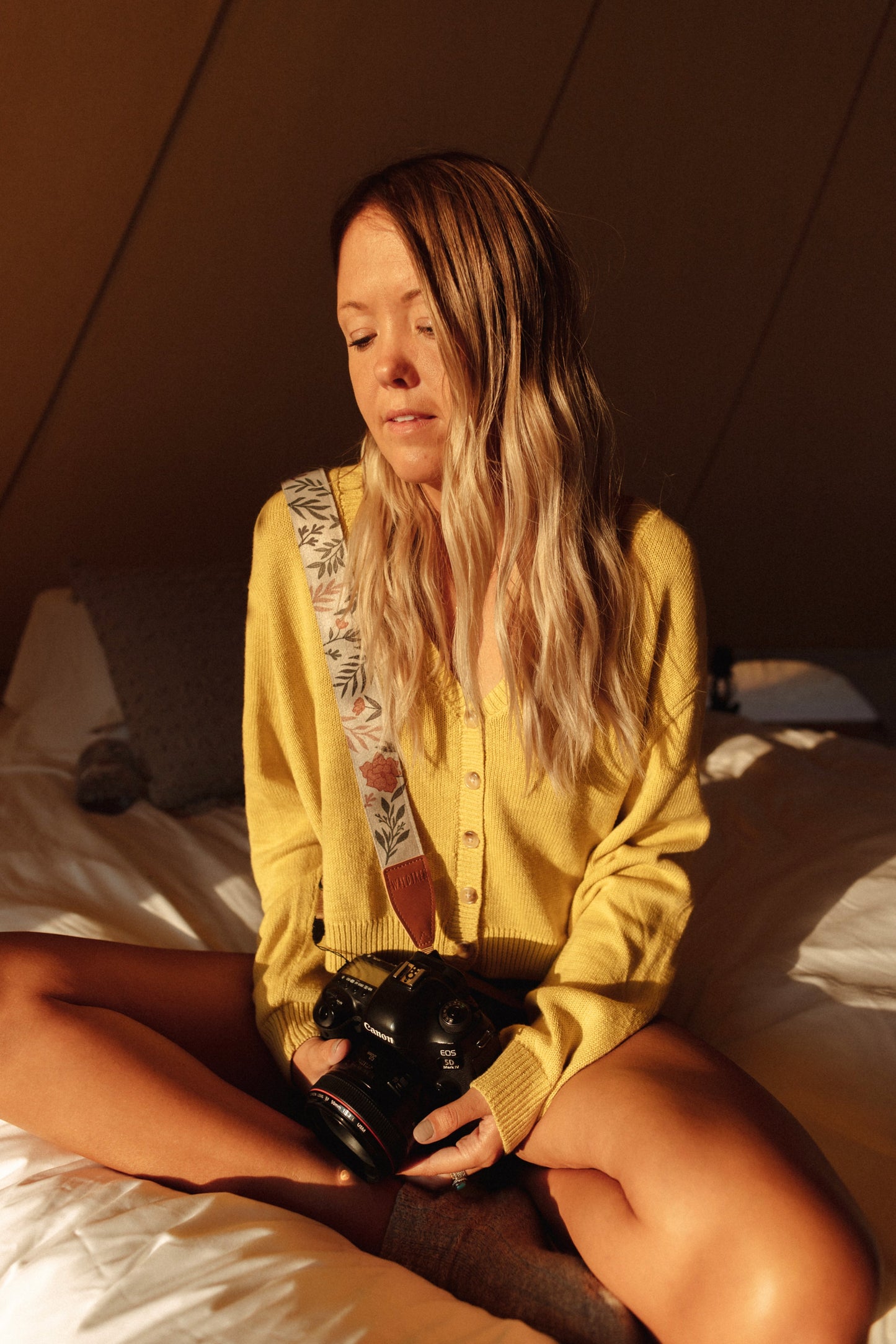 Girl sitting on bed with wildtree moody flower floral print camera strap on shoulder connected to canon camera