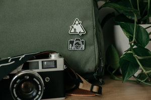 Recycle logo redesigned incorporating a mountain and tree pinned to a backpack above a camera pin and camera.