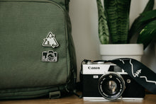 Load image into Gallery viewer, Two Wildtree pins attached to backpack next to film camera and plant
