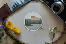 Load image into Gallery viewer, Wildtree Sunrise Lake Sticker featuring lake, mountains, trees and sun sitting on wood surrounded by camera, flowers, and strap
