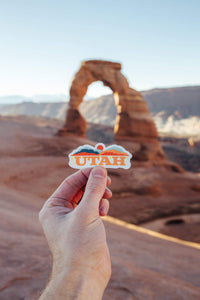 Hand holding Utah sticker in front of Delicate Arch