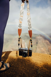 Women holding flower camera strap to side overlooking canyon with waterfall and rainbow