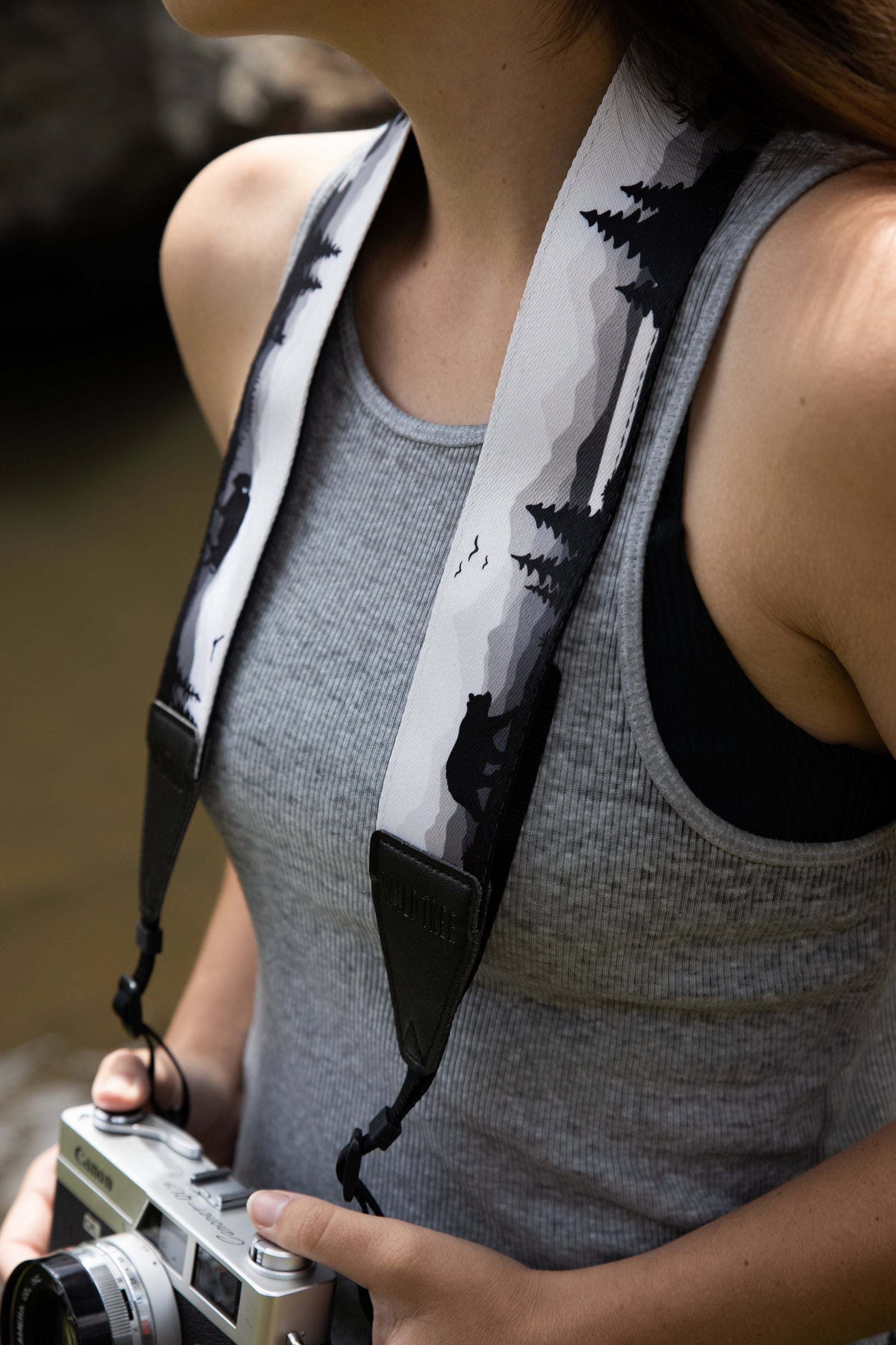 Women wearing Wildtree Wildlife Camera Strap featuring Bear, Moose and Trees with mountain range background attached to Canon film camera