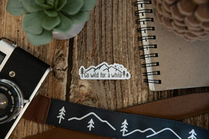 Wildtree go wild for a while sticker on wood background surrounded by notebook, camera and succulent