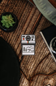 Wildtree Outdoor Photographer Polaroid sticker sitting on wood floor surrounded by camera strap, plants and hat